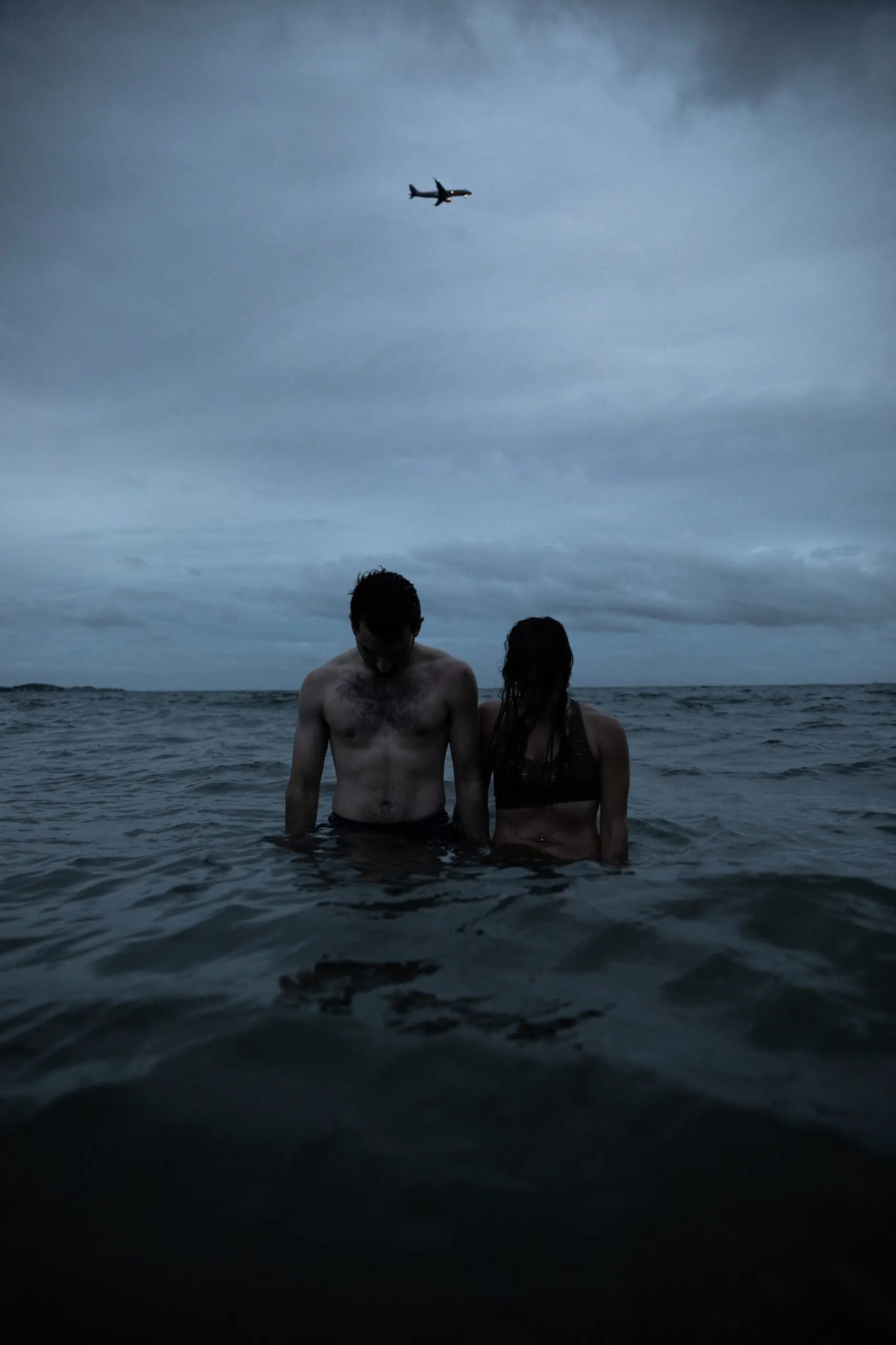 To represent a BPD and Narcissist Couple, this photo is of a woman and man standing in dark water at the beach with a dark gray sky. they're looking down in sadness.